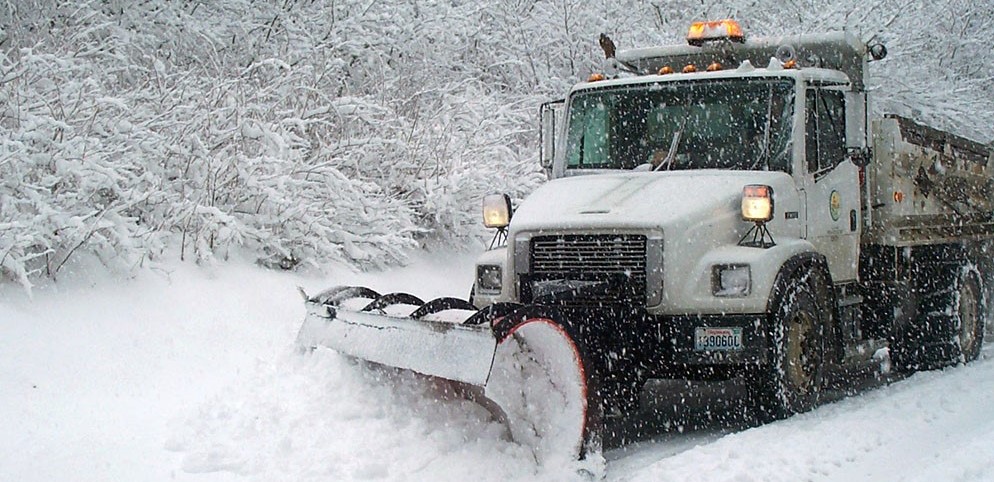 A dump truck-style truck with an orange flashing light and a plow attached pushes snow off a road. Snow is spraying up from the plow. Snow covered branches are in the background.