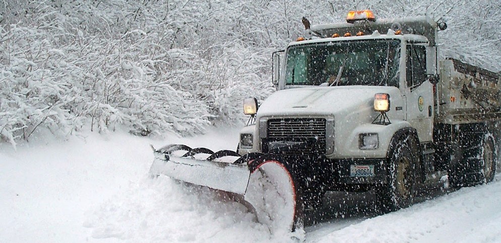 Plow, cropped A dump truck-style truck with an orange flashing light and a plow attached pushes snow off a road. Snow is spraying up from the plow. Snow covered branches are in the background.