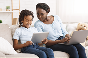 Black woman and daughter sit on sofa, one holding a tablet and the other a laptop computer.
